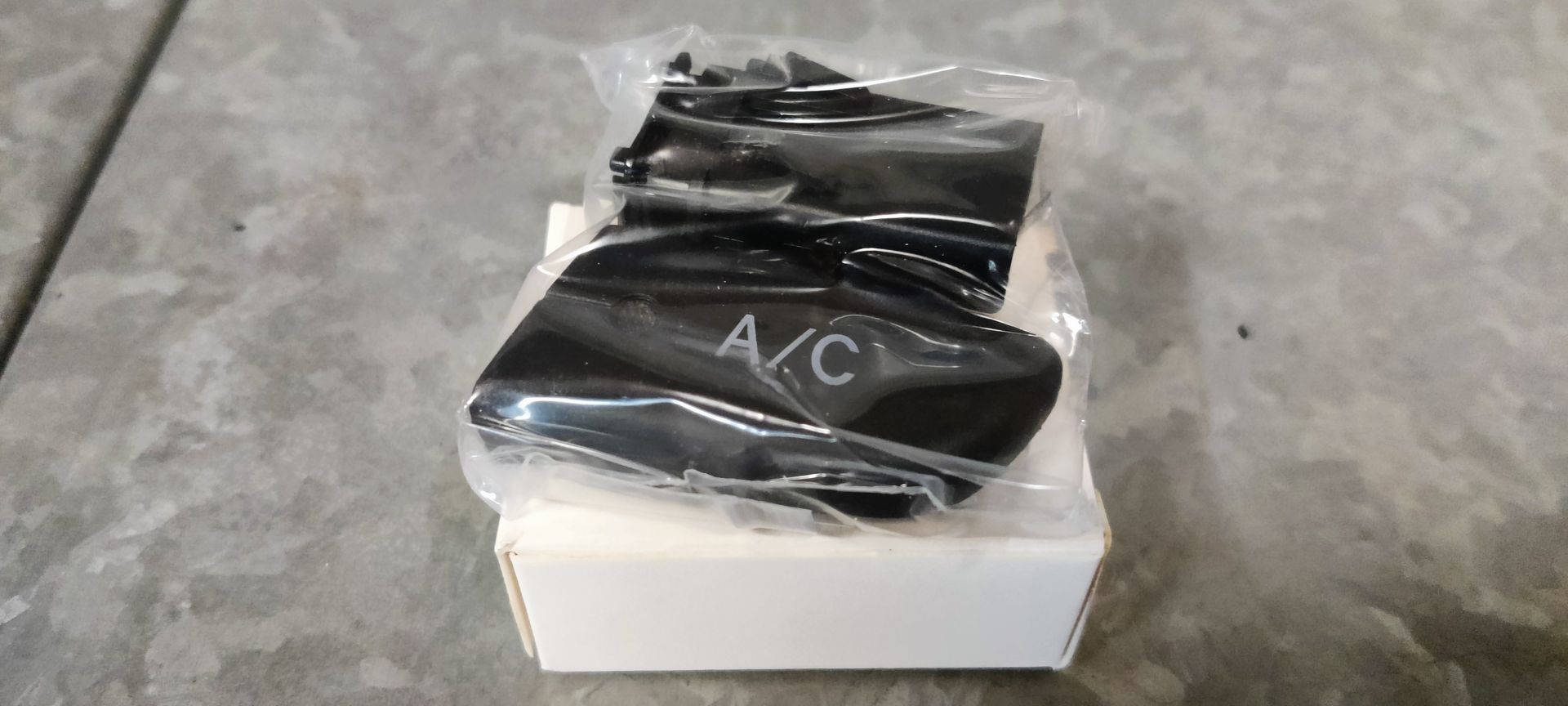 Een klein zwart vervangingsonderdeel voor een knop van de auto-airconditioning, verpakt in een doorzichtig plastic zakje en rustend op een witte doos.