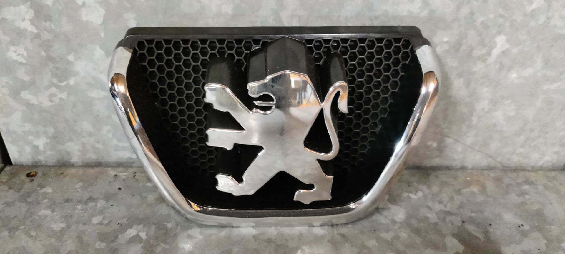 Een verchroomd Peugeot-leeuwenembleem, gemonteerd op een zwart honingraatvormig grilleframe, geplaatst tegen een gestructureerde grijze achtergrond.