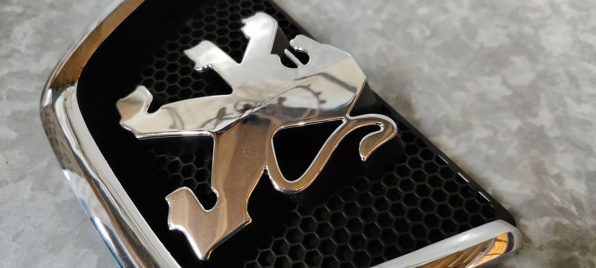 Een verchroomd Peugeot-leeuwenembleem, gemonteerd op een zwart honingraatvormig grilleframe, geplaatst tegen een gestructureerde grijze achtergrond.