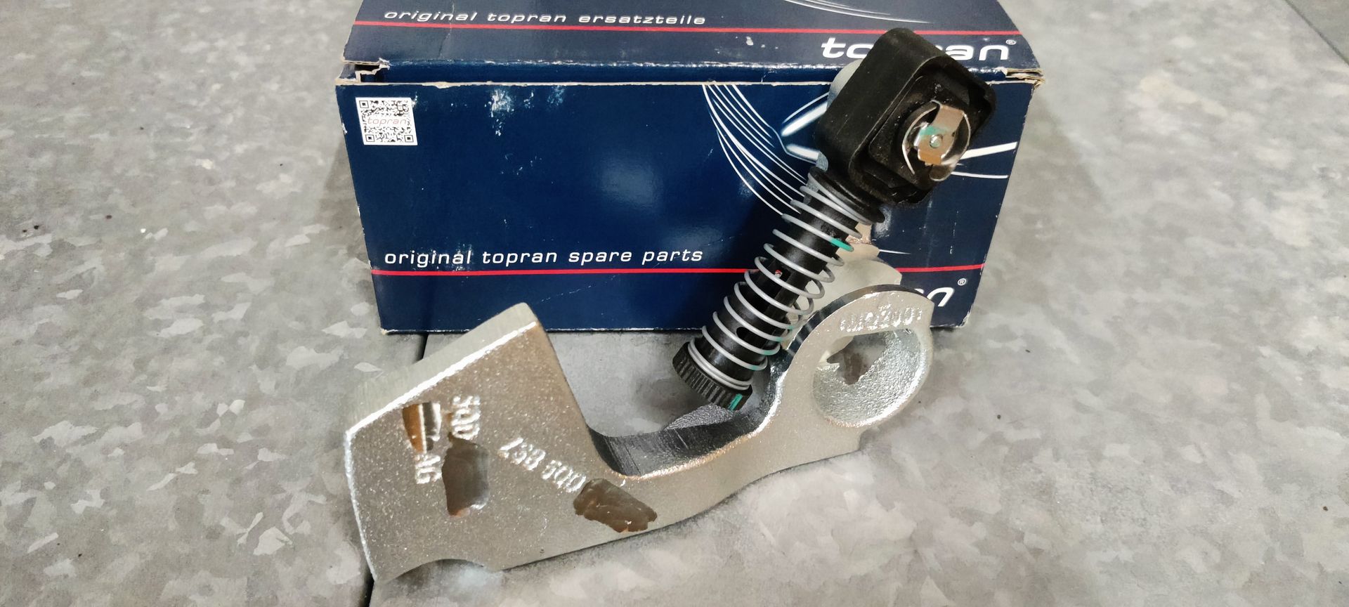 Een metalen spanarm voor auto's met een spiraalveer, geplaatst voor de originele blauwe doos van het merk Toptul.