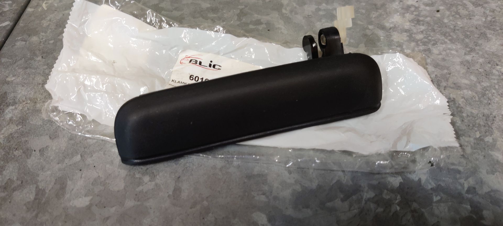 Een matzwarte buitendeurhendel van een auto, rustend op de plastic verpakking tegen een grijze, gestructureerde ondergrond.