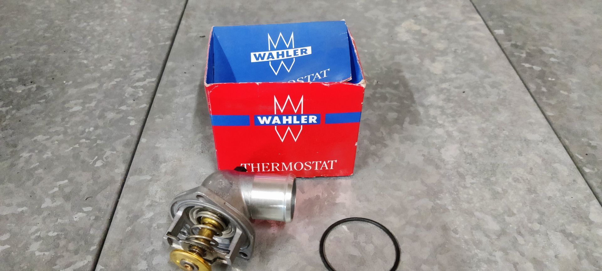 Een thermostaat van het merk Wahler, de rode en blauwe verpakking ervan, en een rubberen O-ringafdichting, tentoongesteld op een tegelvloer.