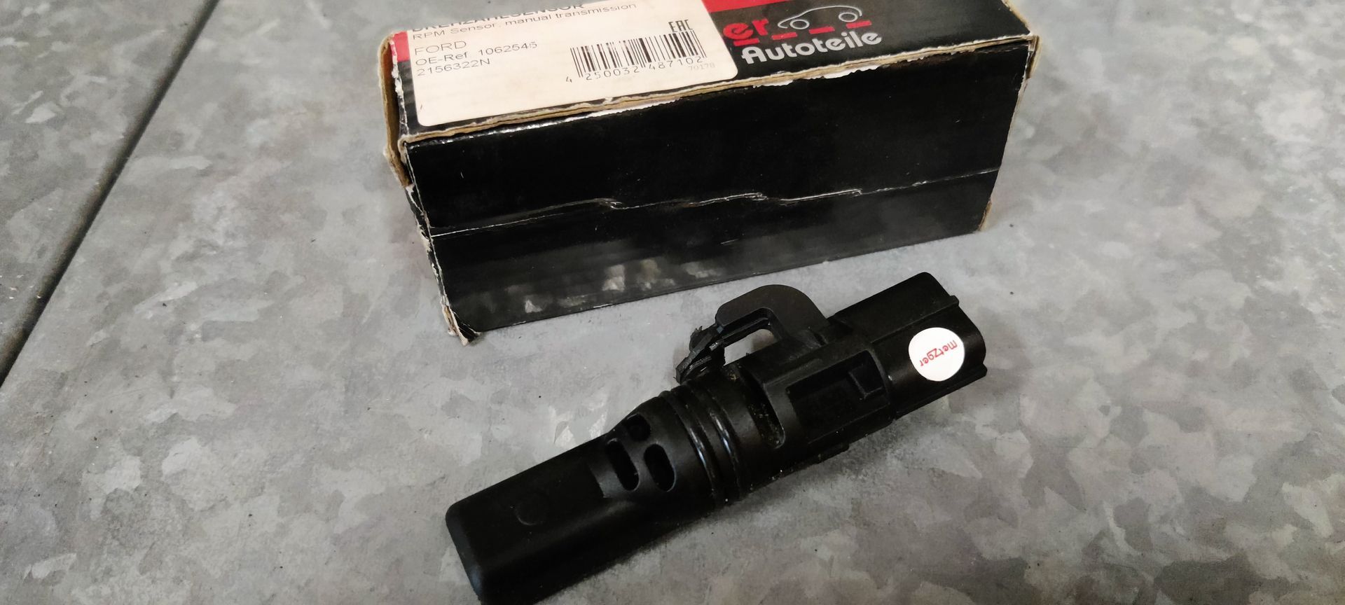 Een zwarte, buisvormige plastic autosensor ligt op een grijze ondergrond voor de originele zwart-witte kartonnen doos.