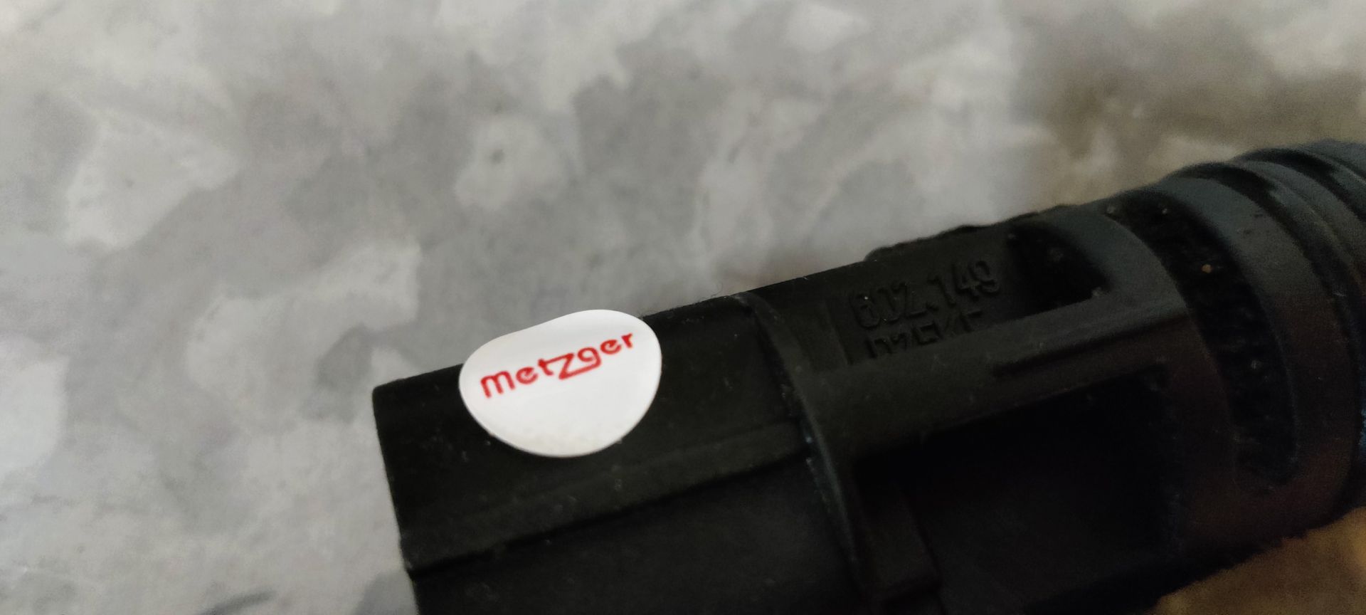 Een zwarte, buisvormige plastic autosensor ligt op een grijze ondergrond voor de originele zwart-witte kartonnen doos.