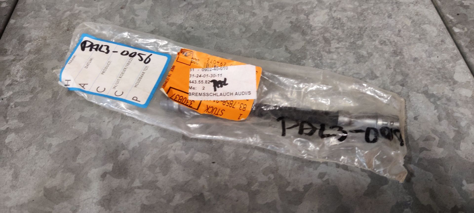 Een metalen industrieel onderdeel in een plastic zak met twee witte labels en handgeschreven etiketten op een metalen oppervlak.