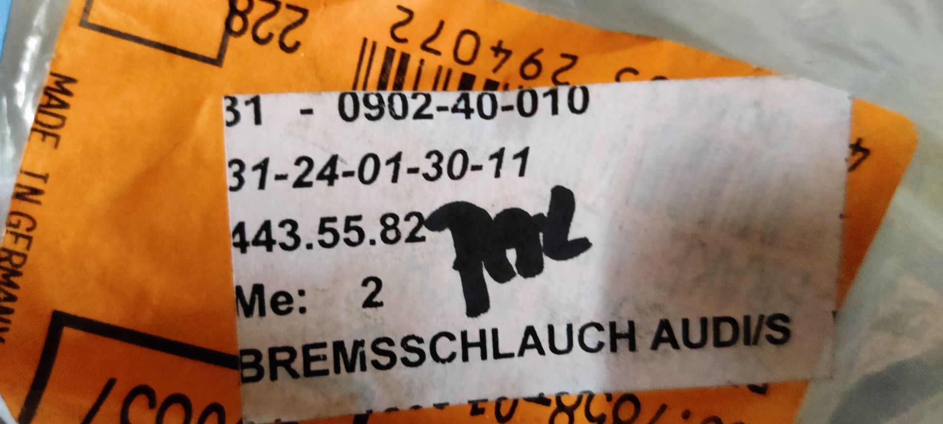 Een metalen industrieel onderdeel in een plastic zak met twee witte labels en handgeschreven etiketten op een metalen oppervlak.