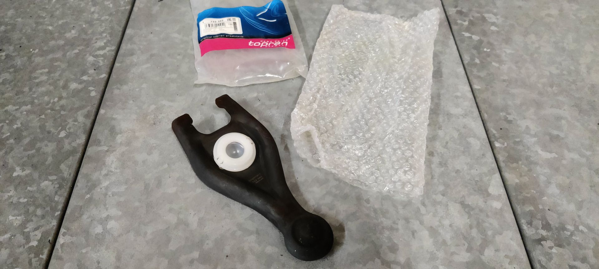 Een zwarte koppelingshendel voor auto's ligt op een tegelvloer naast de verpakking en het bubbeltjesplastic.