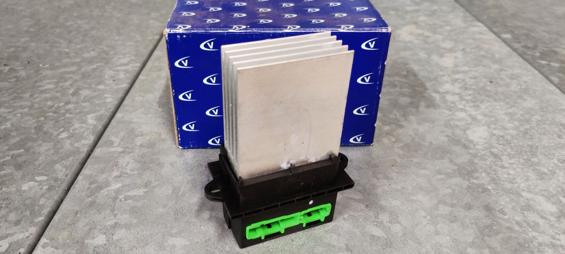 Een weerstand van een ventilatormotor in een voertuig, met een zilverkleurig koelblok en een groene elektrische connector, geplaatst voor een blauwe doos.