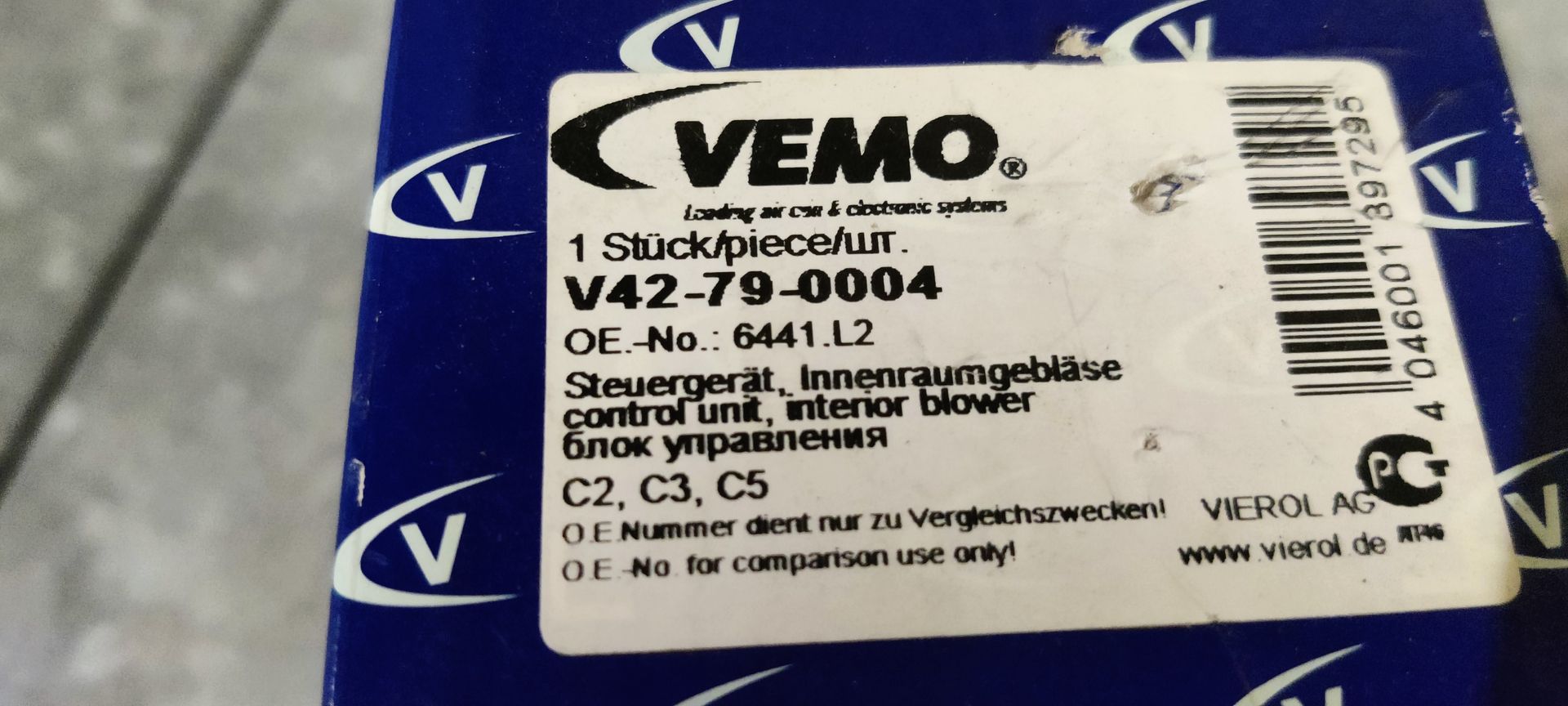 Een weerstand van een ventilatormotor in een voertuig, met een zilverkleurig koelblok en een groene elektrische connector, geplaatst voor een blauwe doos.
