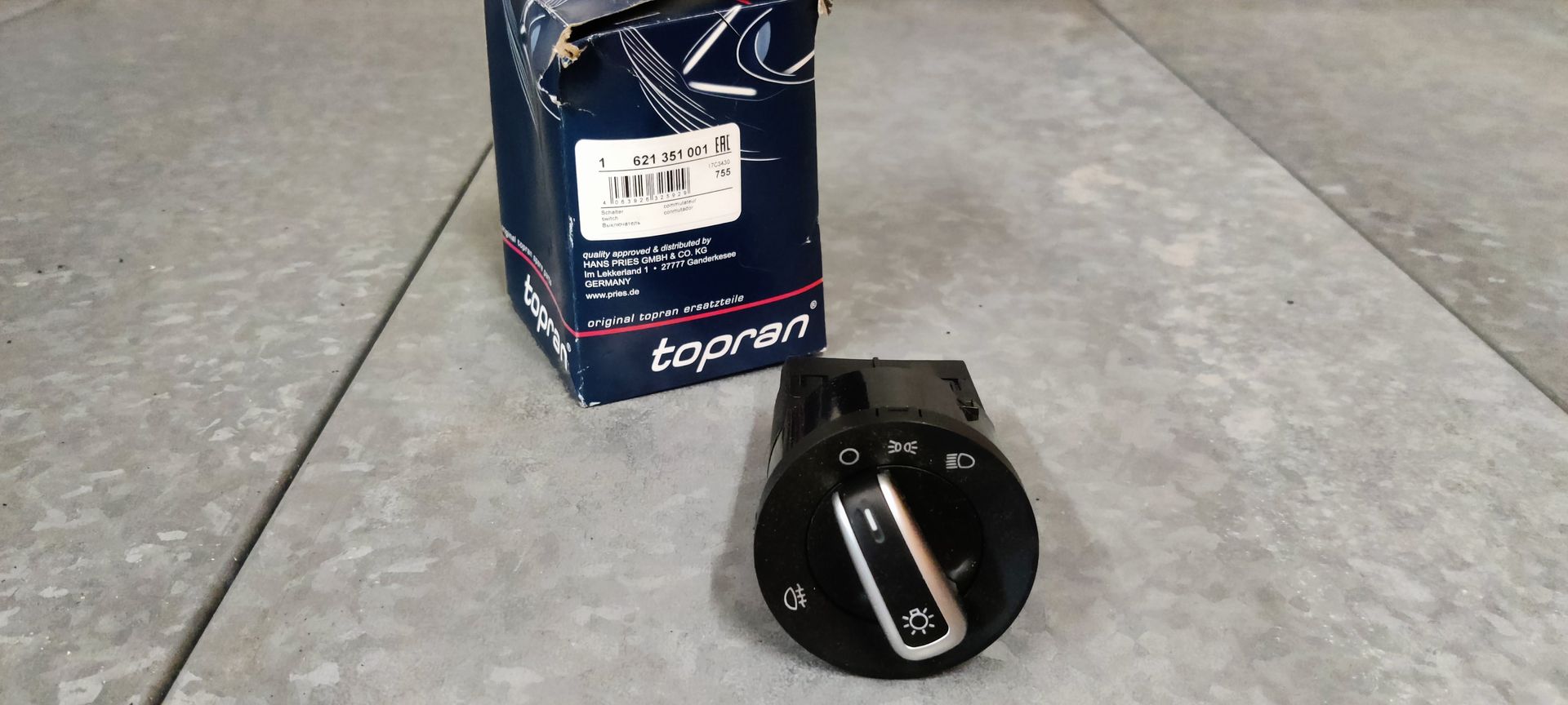 Een Topran-koplampbedieningsschakelaar met een zwarte knop met zilverkleurige accenten, geplaatst op een grijze ondergrond.