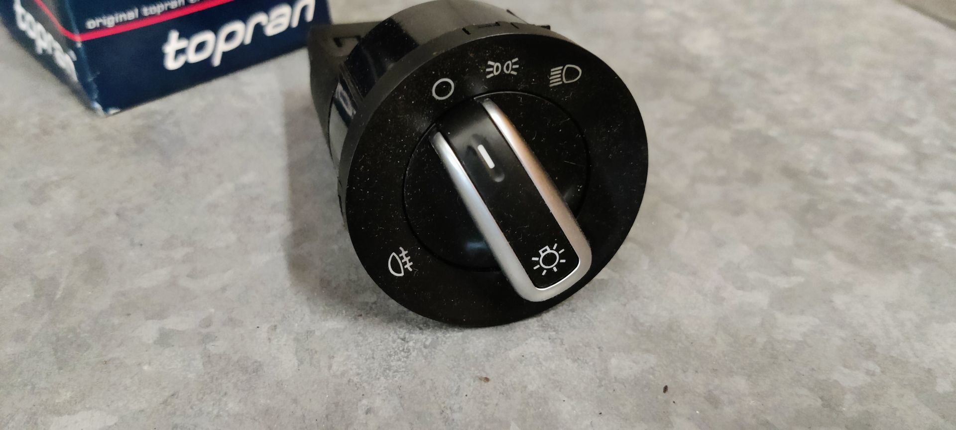 Een Topran-koplampbedieningsschakelaar met een zwarte knop met zilverkleurige accenten, geplaatst op een grijze ondergrond.
