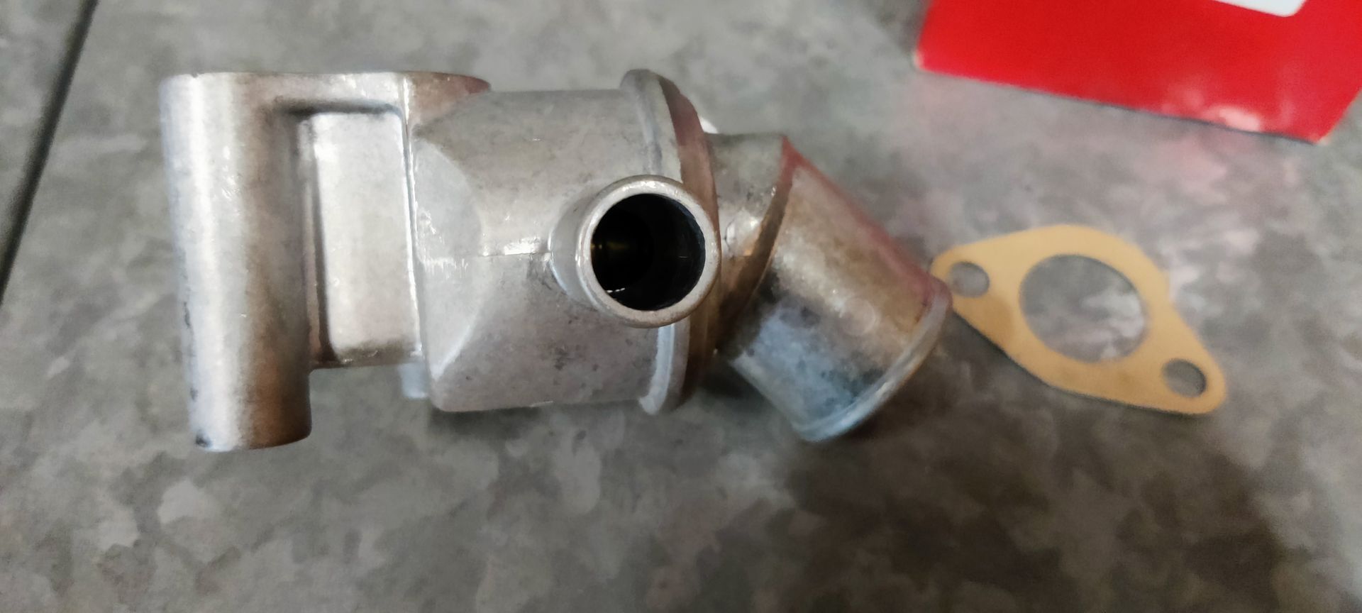 Een metalen thermostaathuis voor auto's met een kleine pijpaansluiting en een bijpassende pakking op een grijs oppervlak.