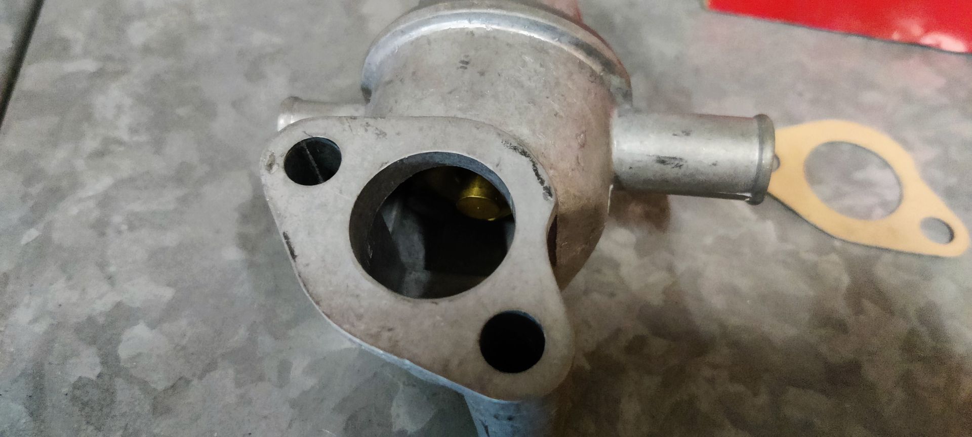 Een metalen thermostaathuis voor auto's met een kleine pijpaansluiting en een bijpassende pakking op een grijs oppervlak.