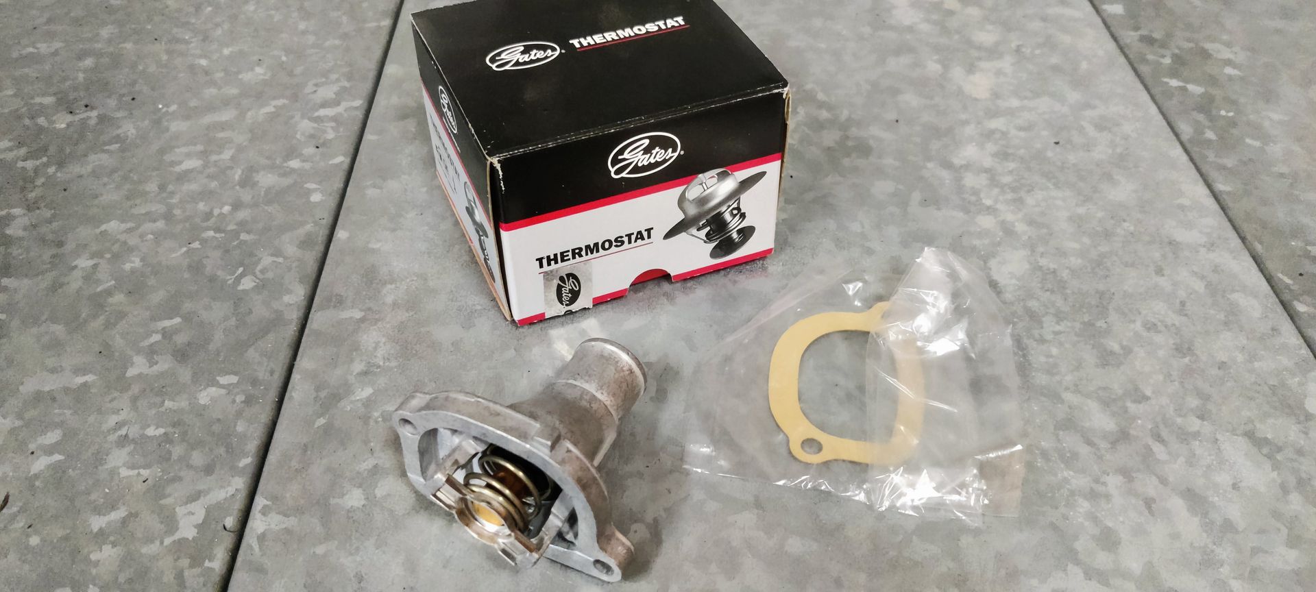 Een thermostaat voor een automotor en een papieren pakking liggen op een grijze vloer naast de zwart-witte verpakking van het product.