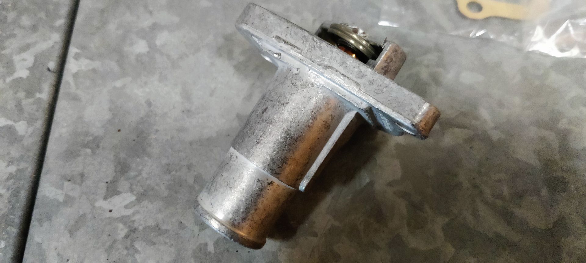 Een thermostaat voor een automotor en een papieren pakking liggen op een grijze vloer naast de zwart-witte verpakking van het product.