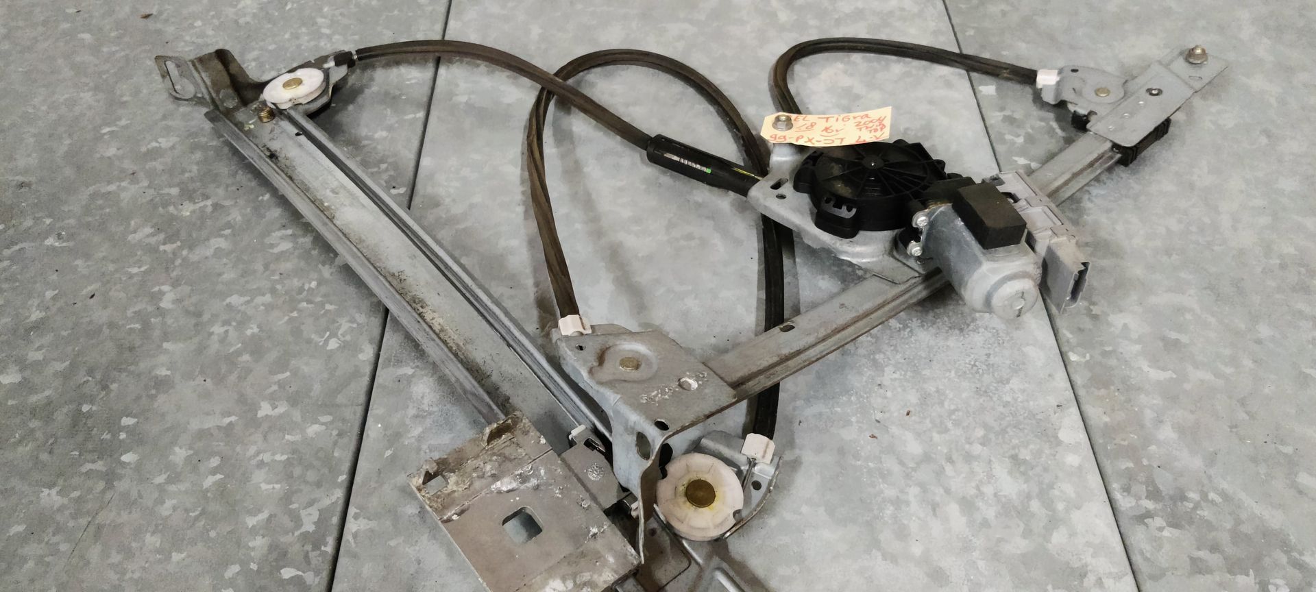 Een metalen raammechanisme voor auto's met een zwarte motor en kabels, geplaatst op een grijze betonnen ondergrond.