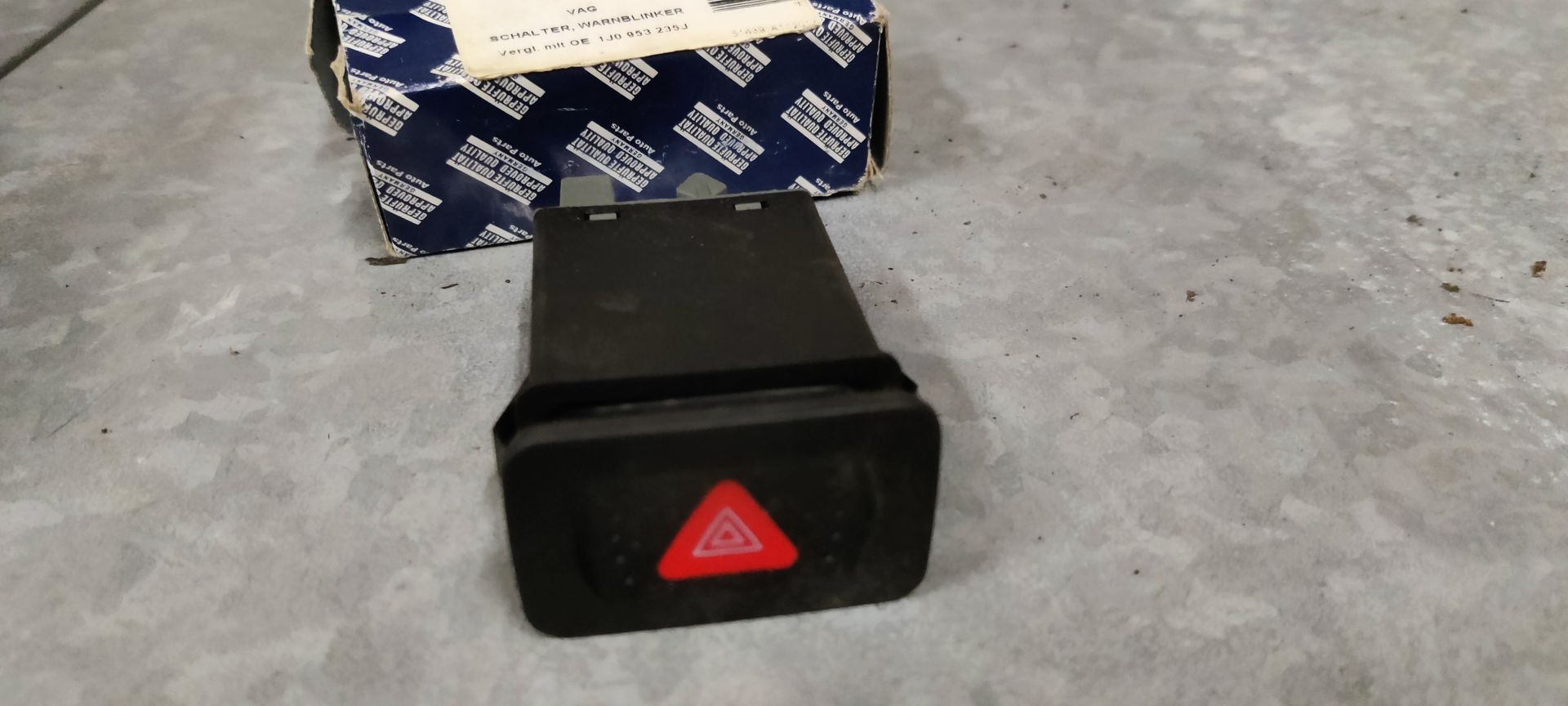 Een zwarte autoalarmlichtschakelaar met een rood driehoekssymbool, die op een grijze ondergrond voor de productverpakking staat.