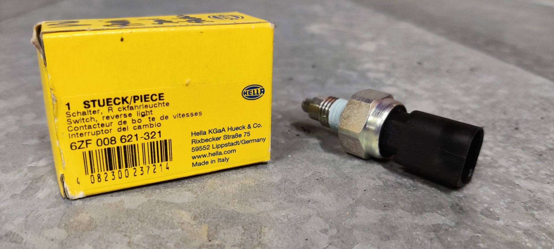 Een Hella-oliedruksensor ligt op een grijze ondergrond naast de gele verpakking met het opschrift onderdeelnummer 6ZF 008 621-321.