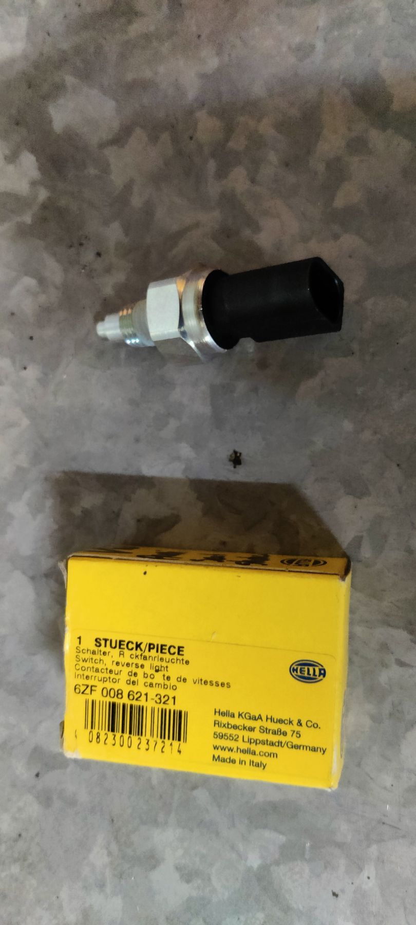 Een Hella-oliedruksensor ligt op een grijze ondergrond naast de gele verpakking met het opschrift onderdeelnummer 6ZF 008 621-321.