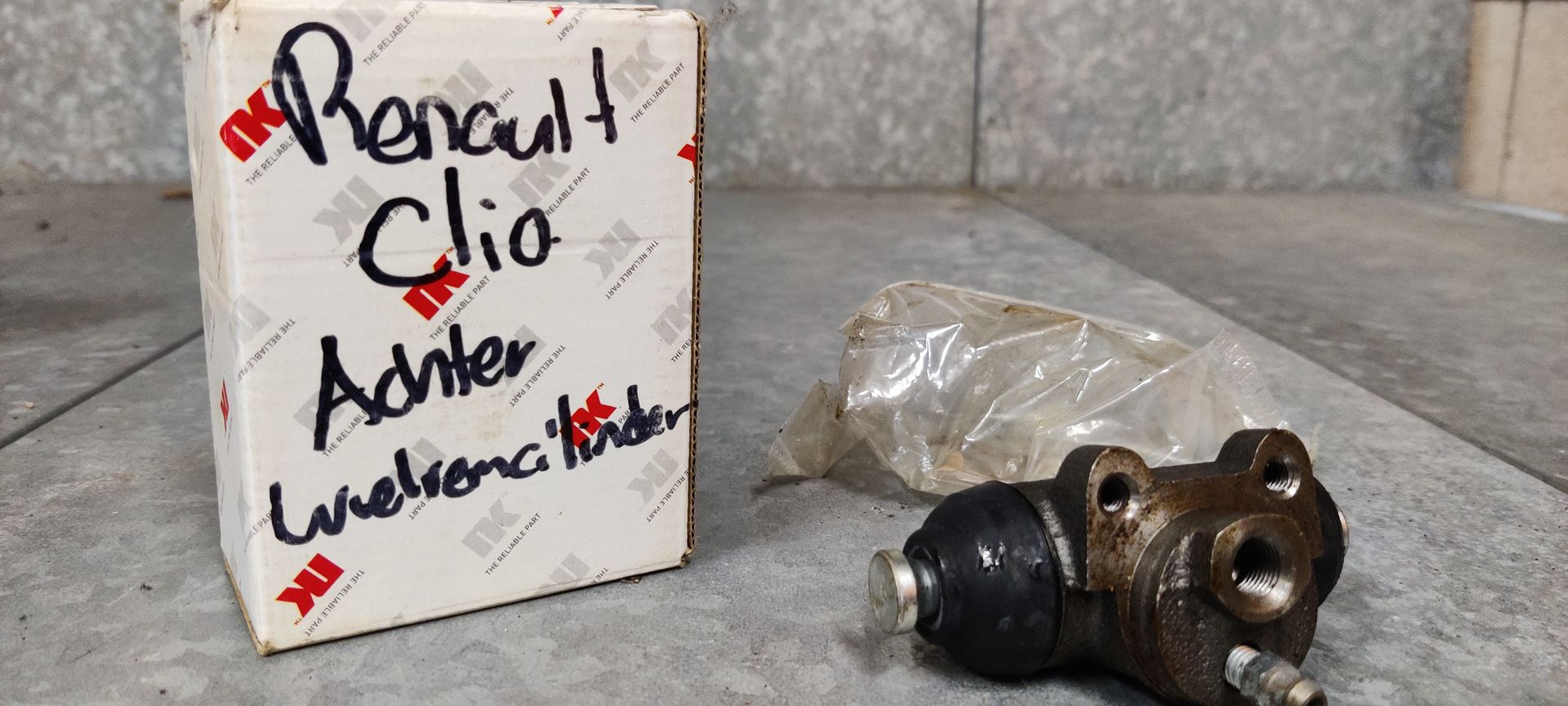 Een metalen wielcilinder voor een Renault Clio, rustend op een grijze ondergrond naast een doos met handgeschreven tekst.