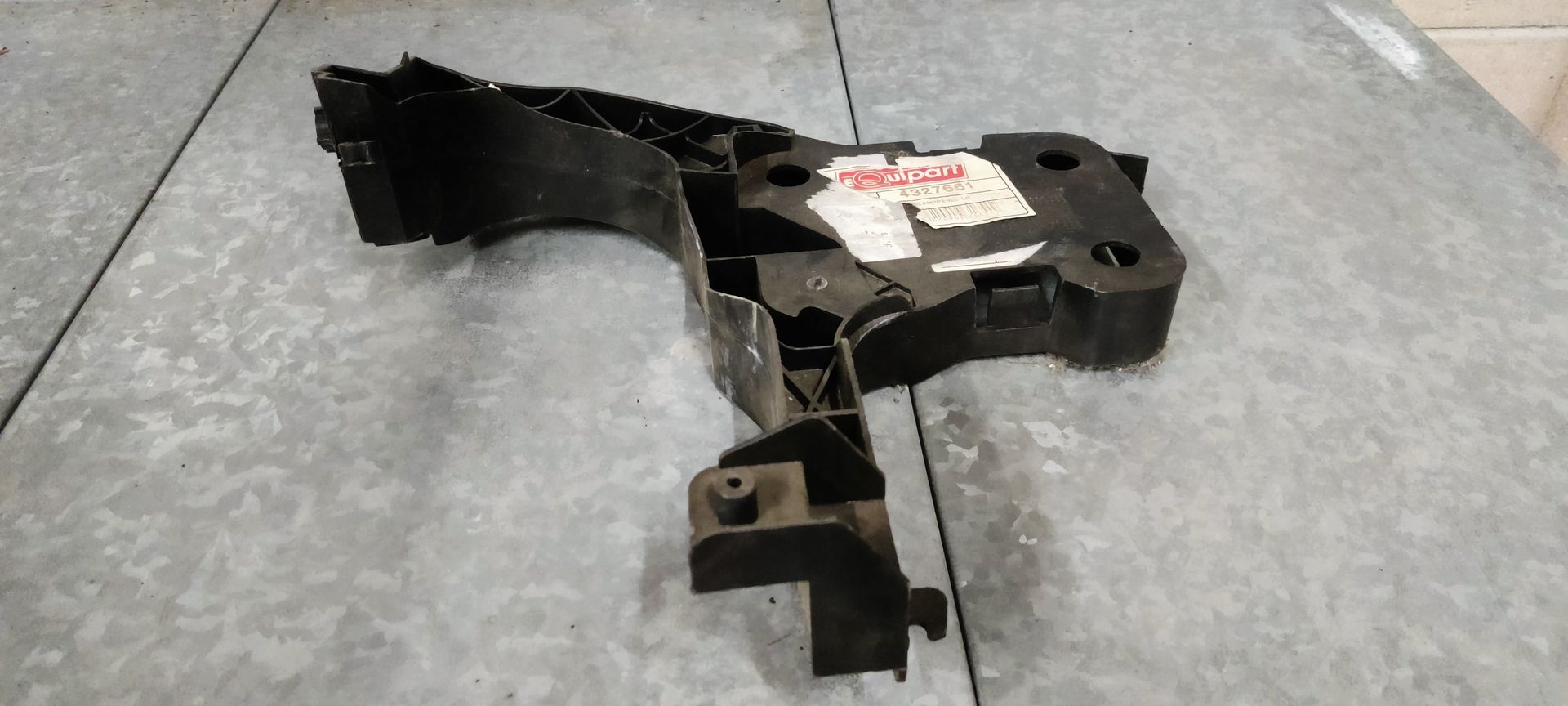 Een zwarte plastic montagebeugel voor autokoplampen met een sticker op een grijze, industriële ondergrond.