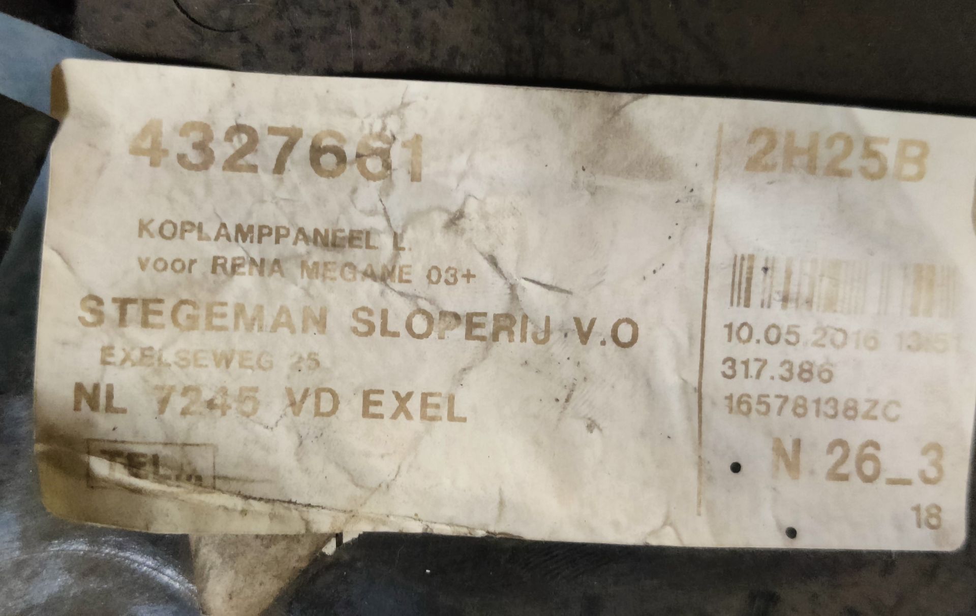 Een zwarte plastic montagebeugel voor autokoplampen met een sticker op een grijze, industriële ondergrond.