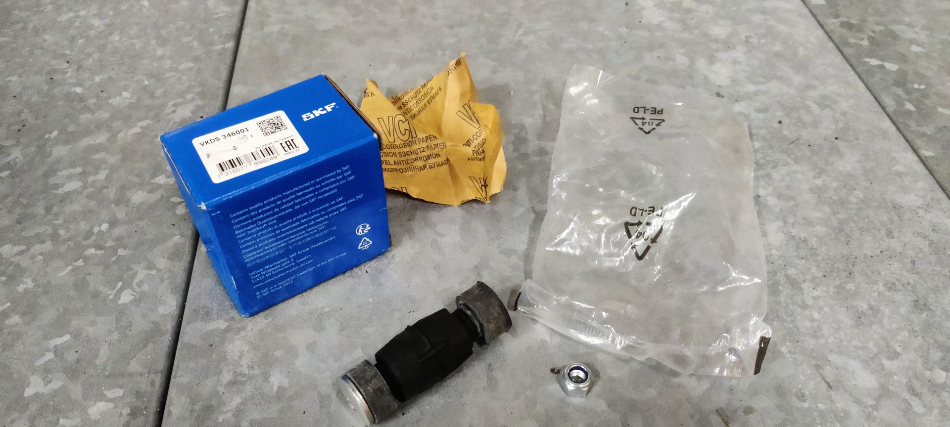 Een blauwe SKF-onderdelendoos, een plastic zak, een klein metalen moertje en een cilindrisch zwart motoronderdeel van rubber en metaal.