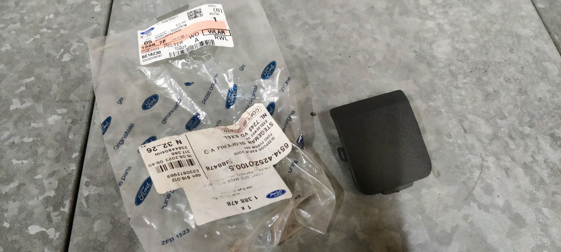 Een plastic zakje met Ford-logo en een etiket ernaast, met daarin een klein, rechthoekig zwart plastic sierstukje voor auto's.