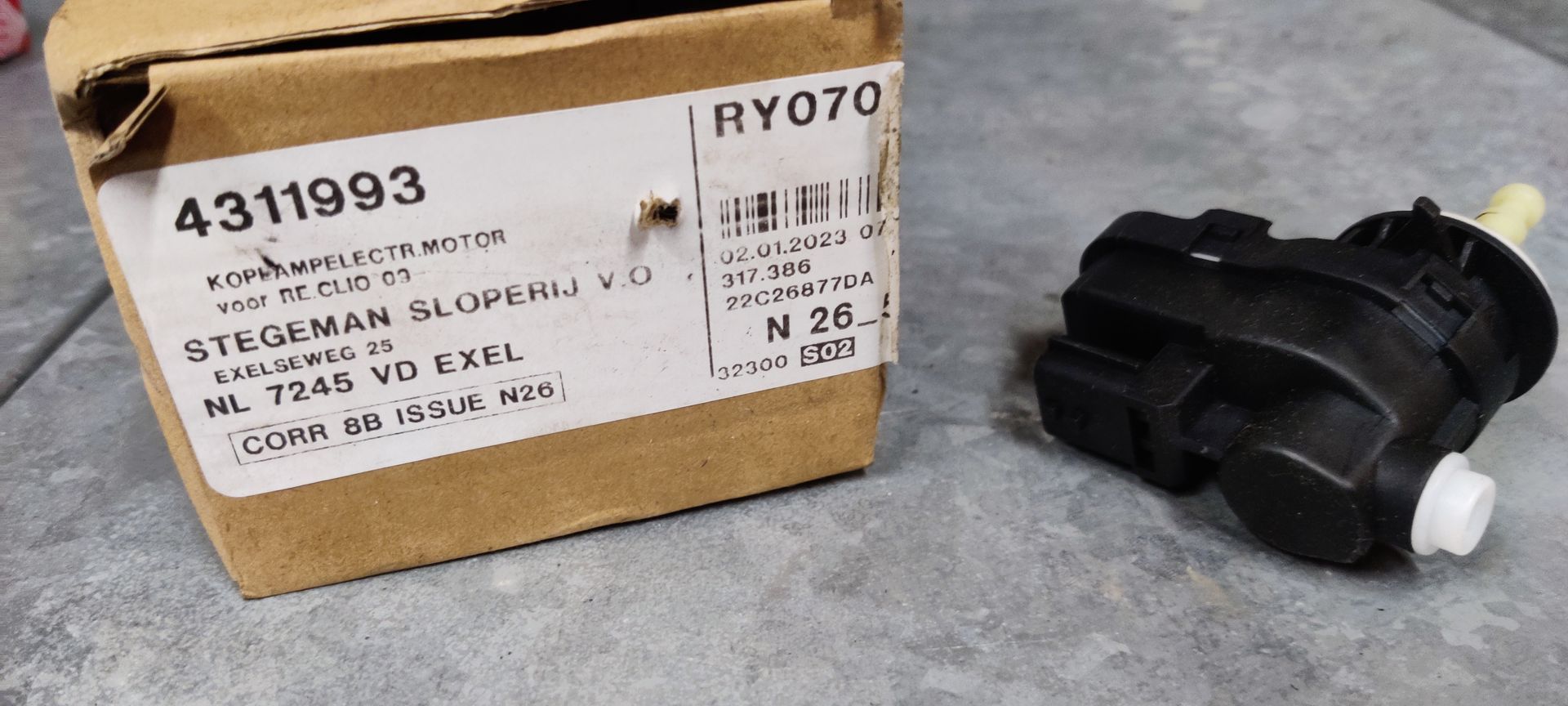 Een nieuw Citroën-onderdeel in een kartonnen doos naast een zwarte elektrische actuatormotor op een grijze ondergrond.