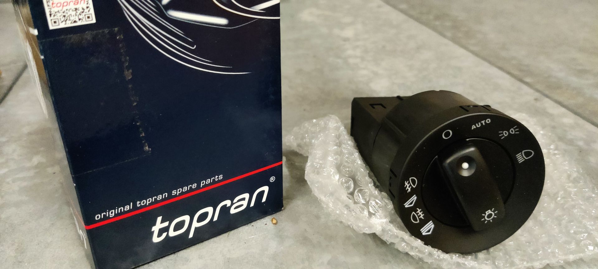 Een doosje van het merk Topran naast een zwarte bedieningsschakelaar voor autokoplampen, geplaatst op een lichtgekleurde ondergrond.