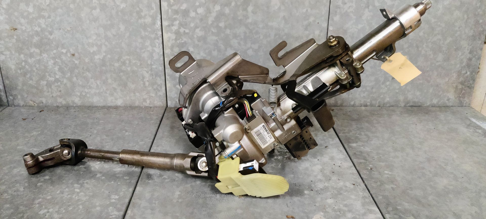 Een metalen stuurkolom voor auto's met bevestigingsbeugels, een motor en een kruiskoppelingsstuk, afgebeeld tegen een grijze muur.