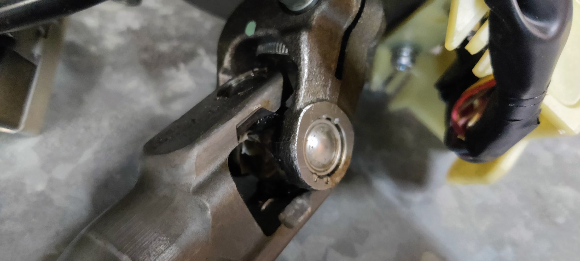 Een metalen stuurkolom voor auto's met bevestigingsbeugels, een motor en een kruiskoppelingsstuk, afgebeeld tegen een grijze muur.