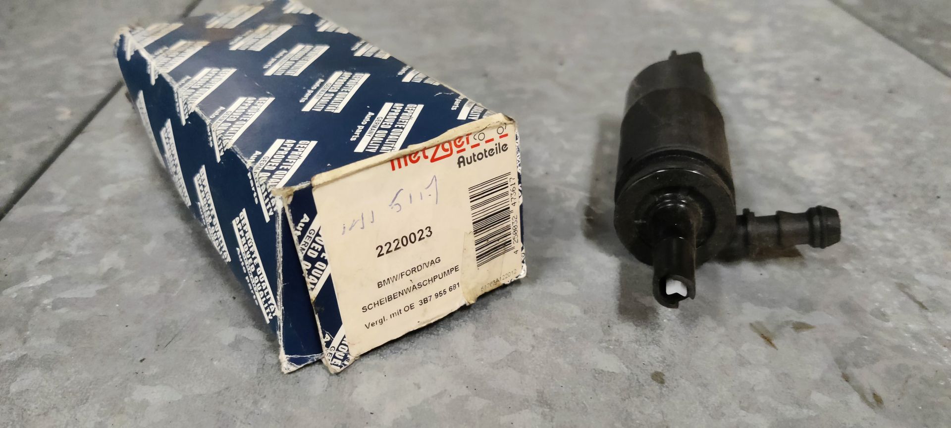 Een zwarte ruitenwisservloeistofpomp voor auto's staat naast de blauw-witte kartonnen verpakking met het merklogo op een grijze vloer.