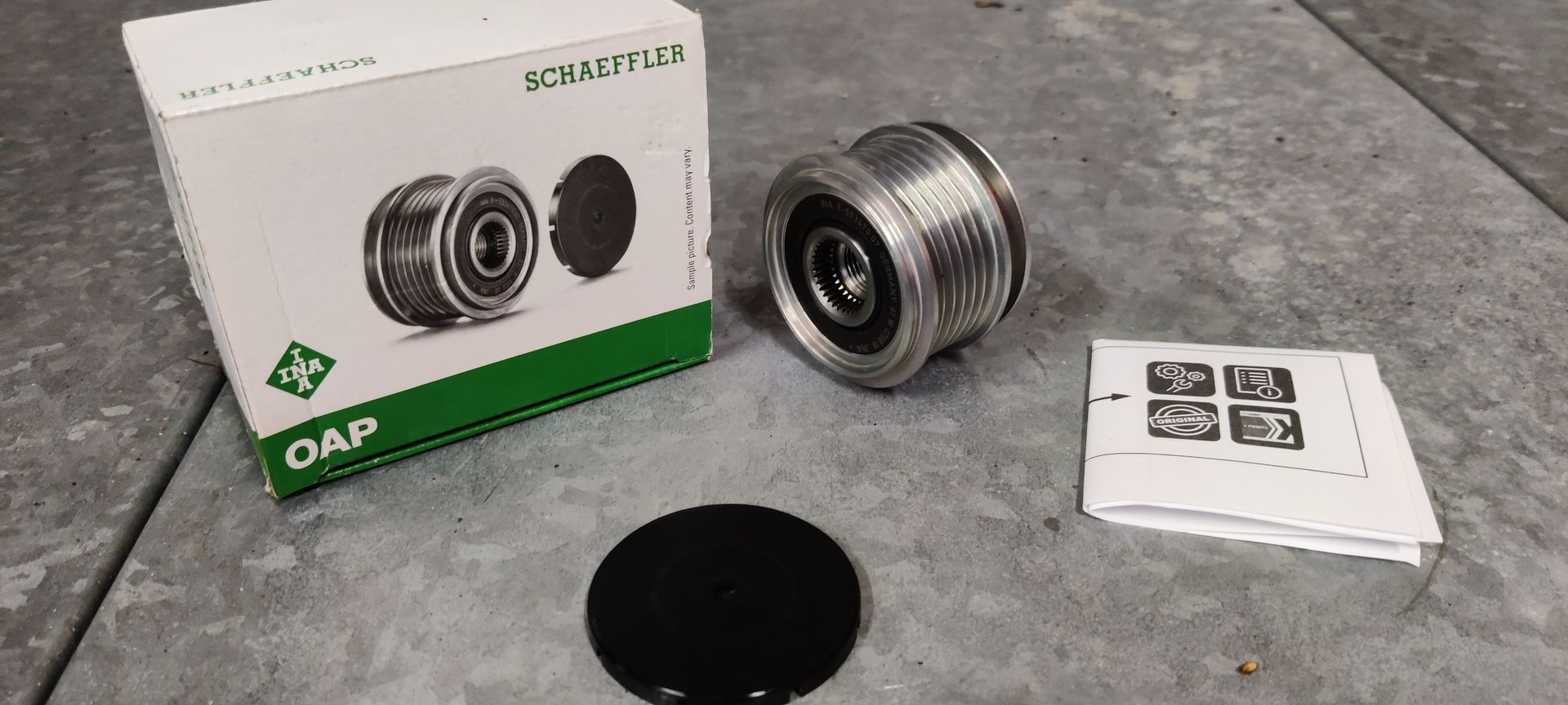 Een Schaeffler OAP-dynamo-poelie in de doos, met beschermkap en handleiding, op een betonnen ondergrond.