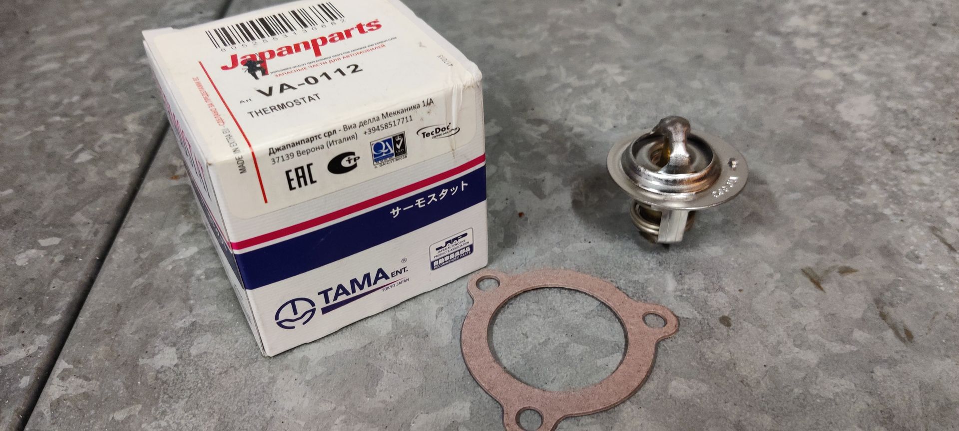 Een thermostaat van het merk Japanparts met bijbehorende papieren pakking, geplaatst op een grijs, gestructureerd oppervlak.