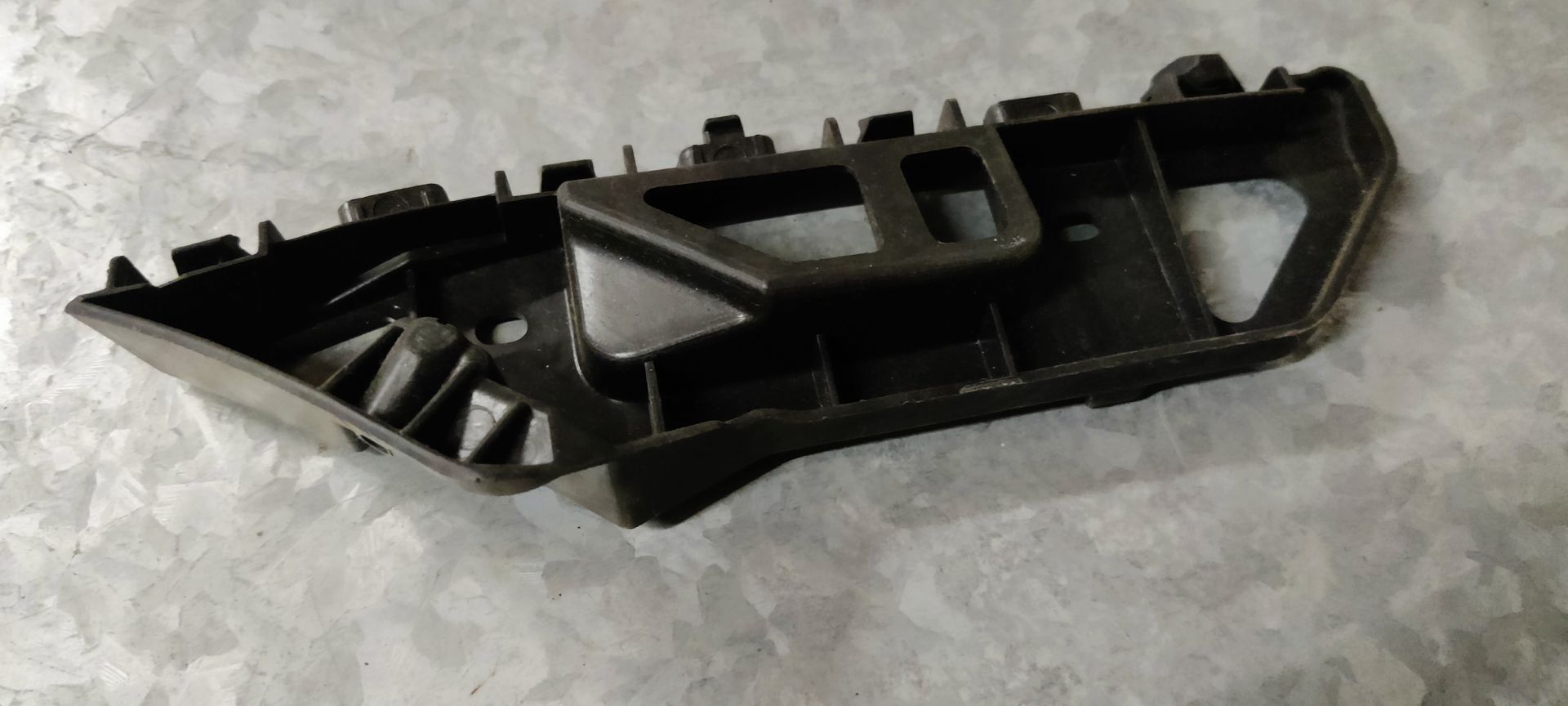 Een zwarte plastic steunbeugel voor een autobumper, met daarachter het witte Niparts-verpakkingslabel op een metalen ondergrond.