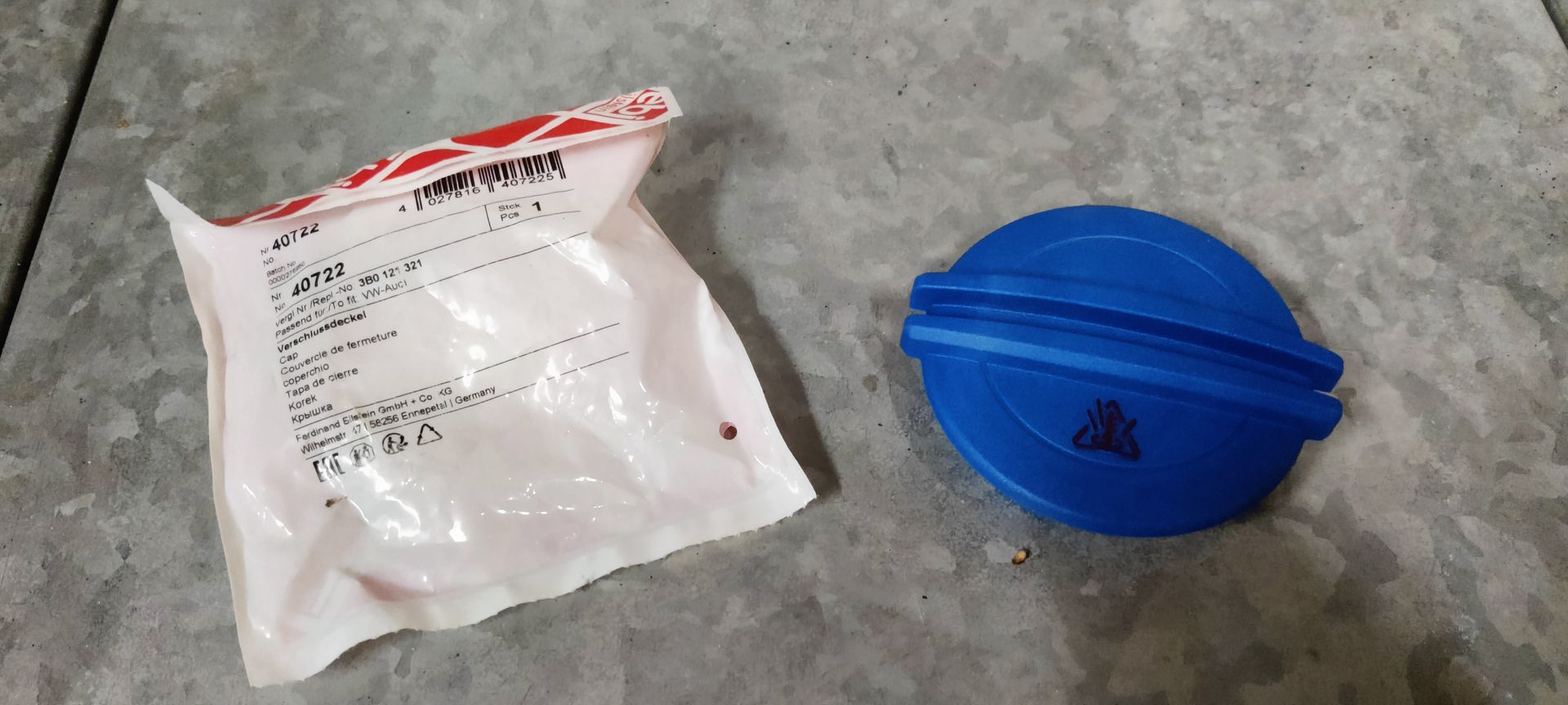 Een blauwe plastic dop van het ruitenwisservloeistofreservoir ligt naast de originele witte plastic verpakking op een grijze vloer.