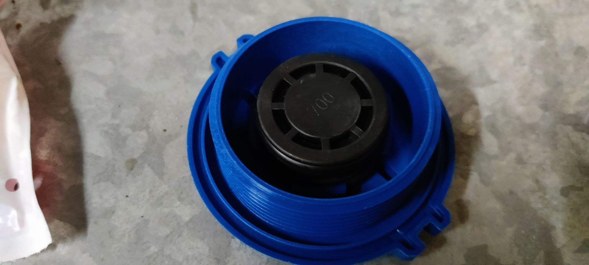 Een blauwe plastic dop van het ruitenwisservloeistofreservoir ligt naast de originele witte plastic verpakking op een grijze vloer.