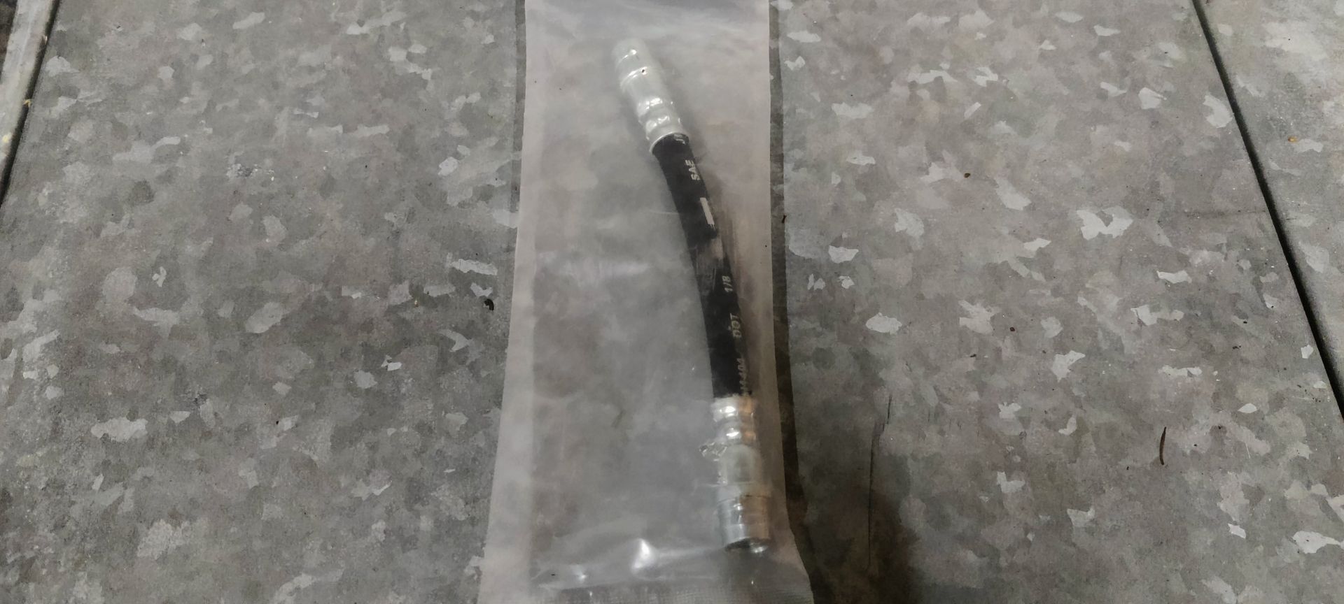 Een korte, zwarte remslang voor auto's met zilverkleurige metalen schroefverbindingen aan beide uiteinden, in een doorzichtige plastic zak.