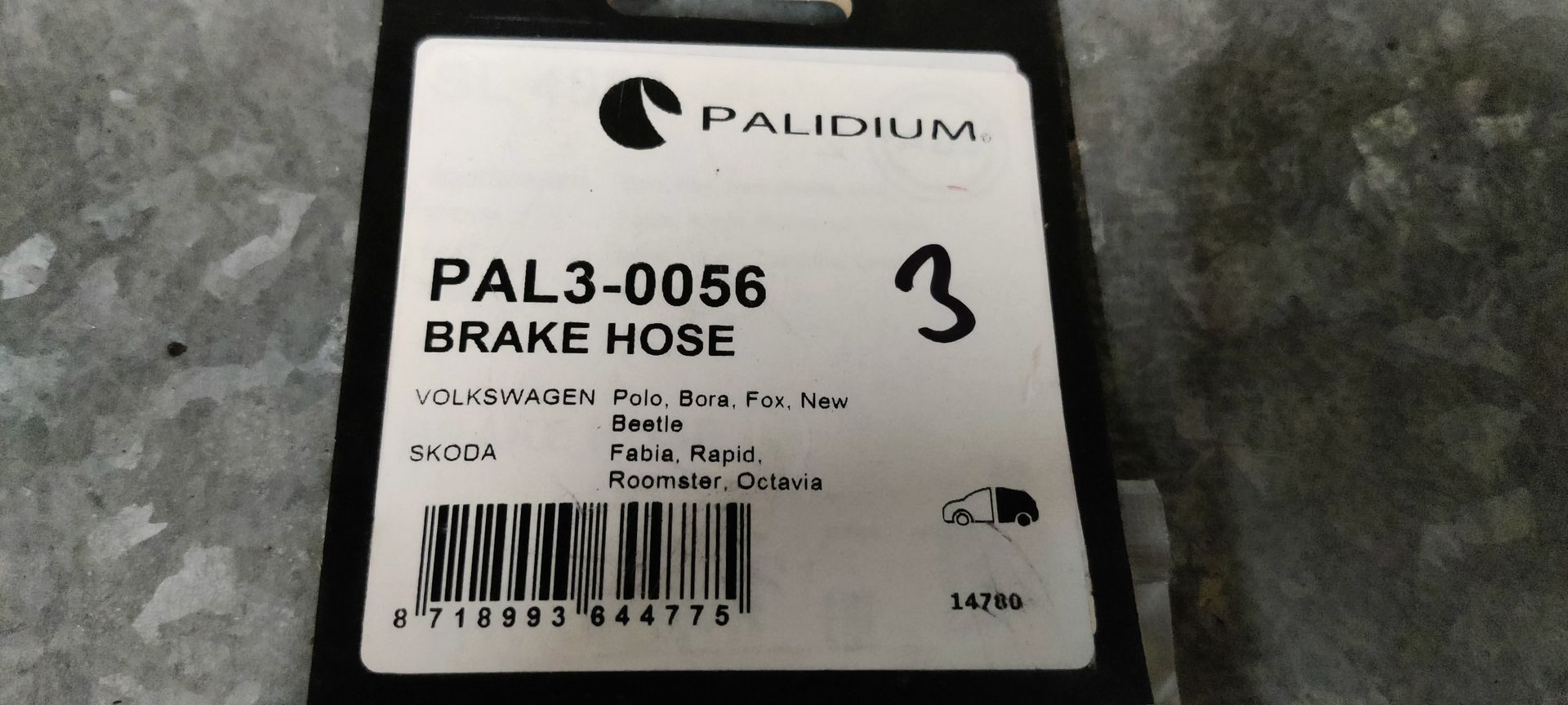 Een korte, zwarte remslang voor auto's met zilverkleurige metalen schroefverbindingen aan beide uiteinden, in een doorzichtige plastic zak.