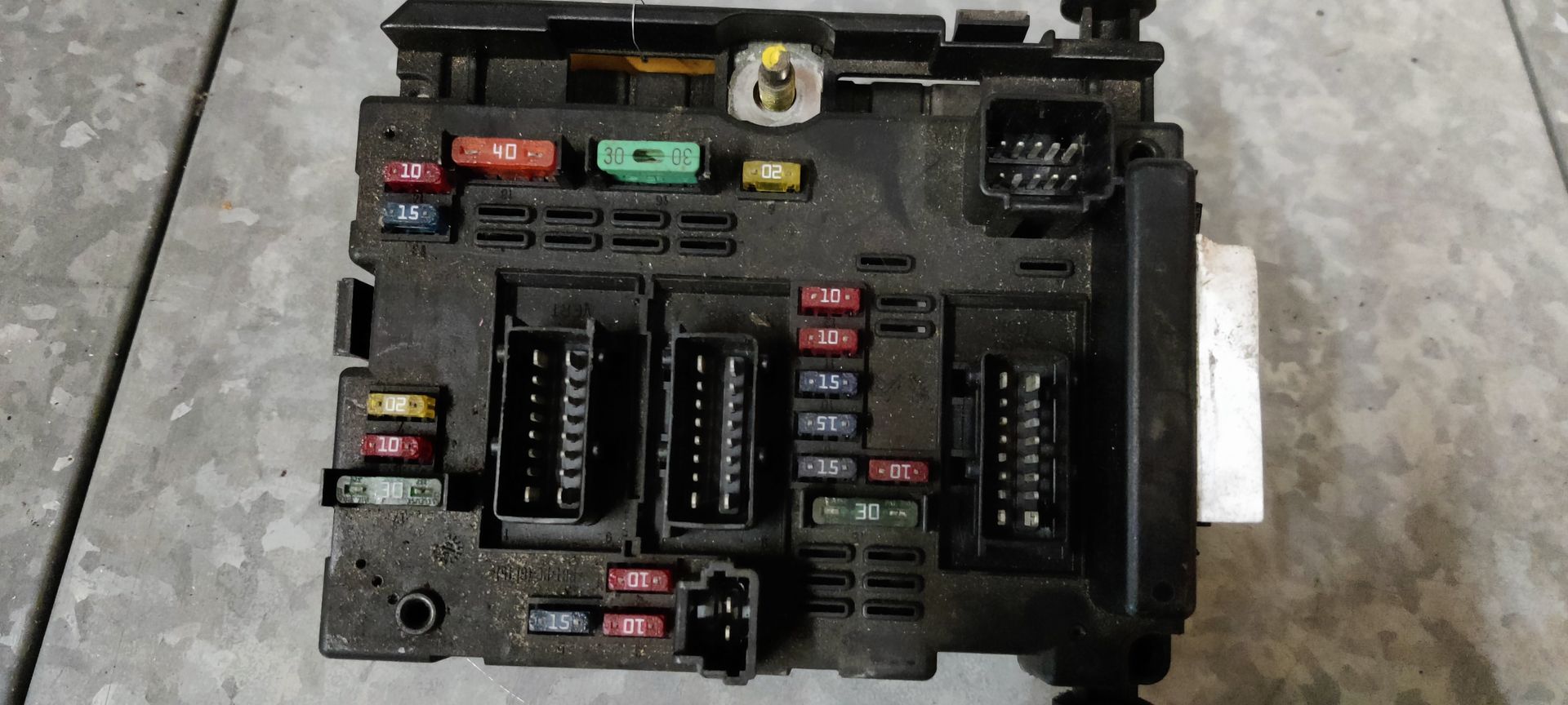 Een zwarte zekeringkast voor auto's met diverse meerkleurige zekeringen en aansluitingen, van bovenaf gezien op een grijze ondergrond.