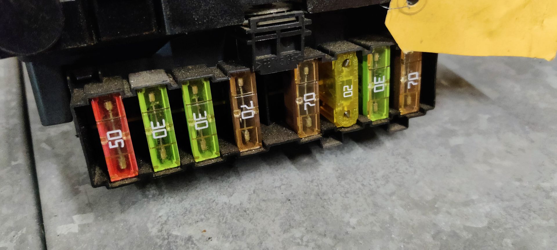 Een zwarte zekeringkast voor auto's met diverse meerkleurige zekeringen en aansluitingen, van bovenaf gezien op een grijze ondergrond.