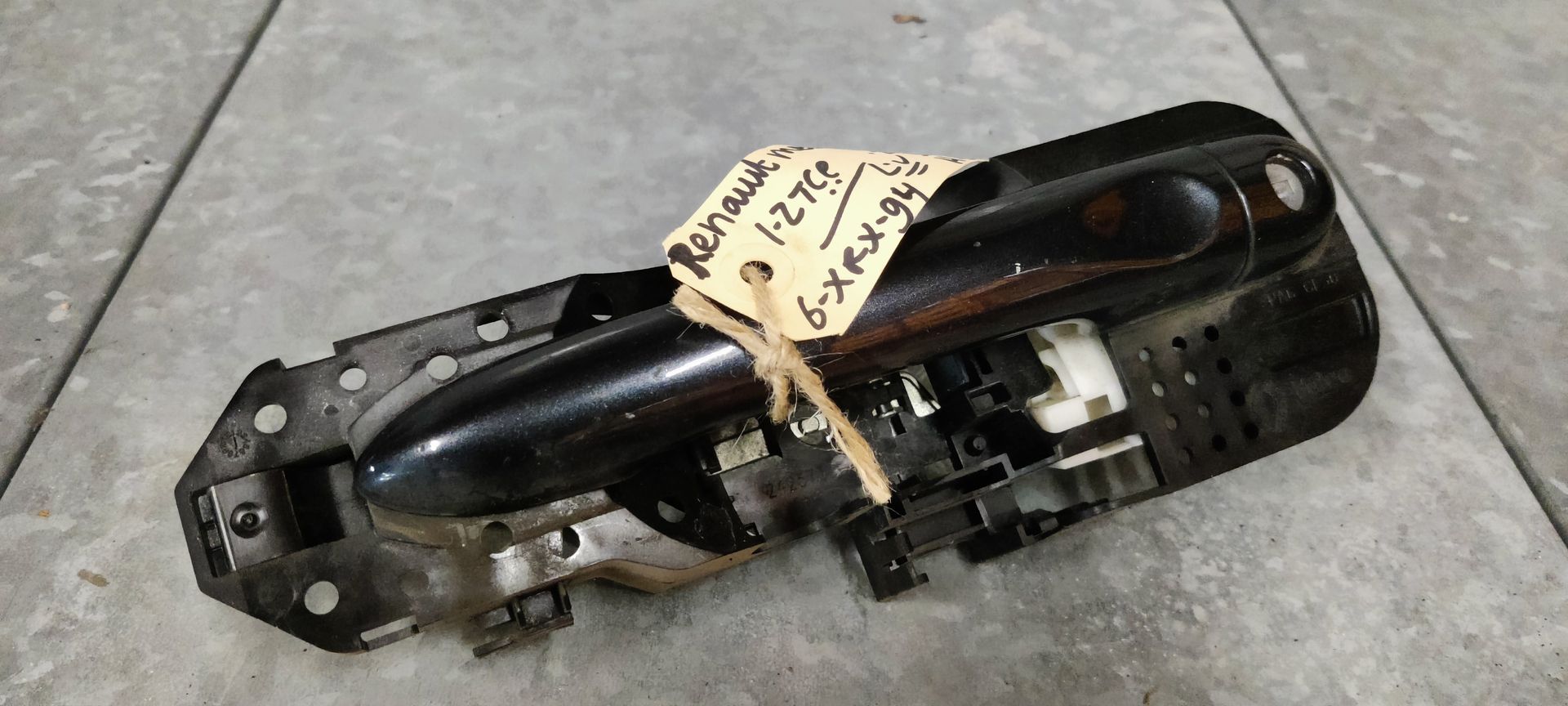 Een zwarte buitendeurhendel van een auto staat op een grijze ondergrond met een handgeschreven identificatielabel dat met een touwtje is bevestigd.