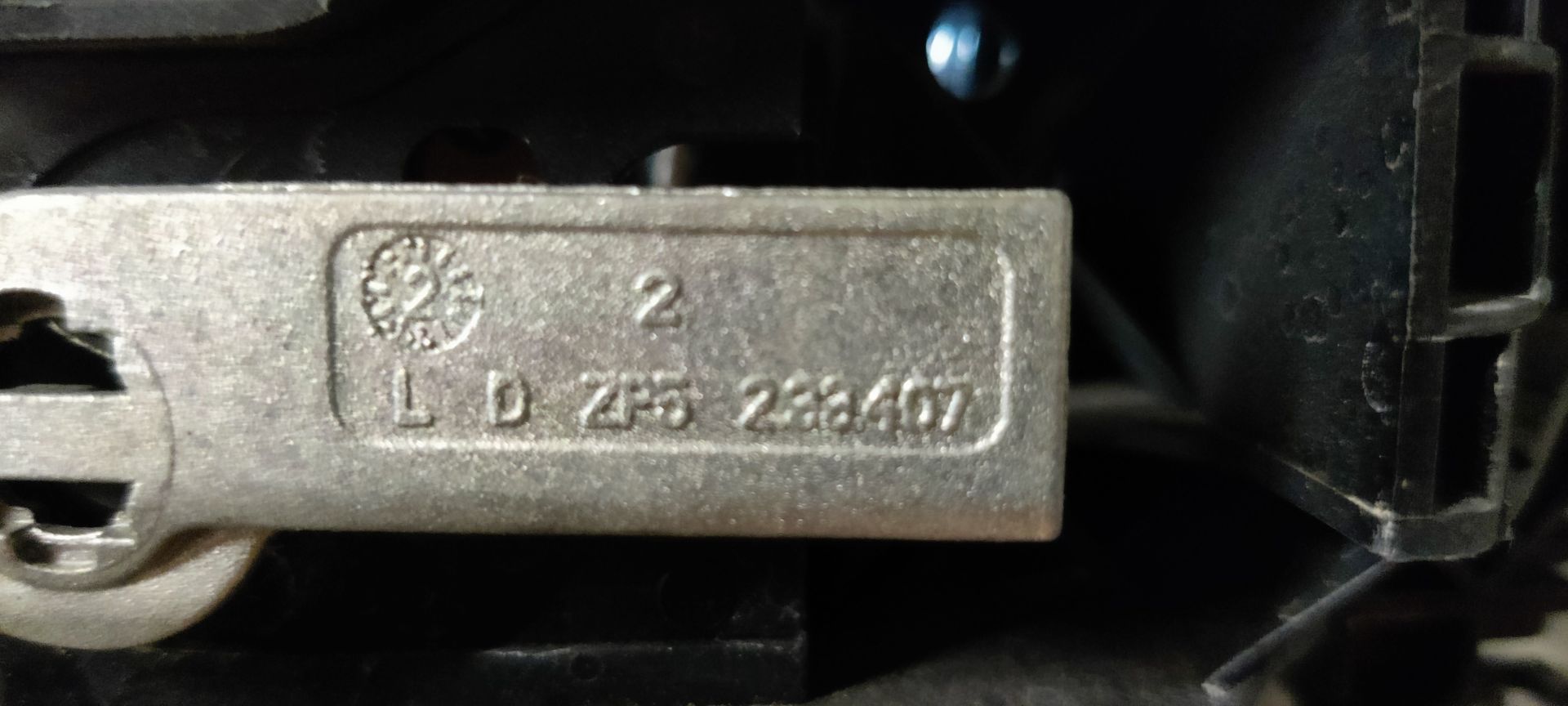 Een zwarte buitendeurhendel van een auto staat op een grijze ondergrond met een handgeschreven identificatielabel dat met een touwtje is bevestigd.