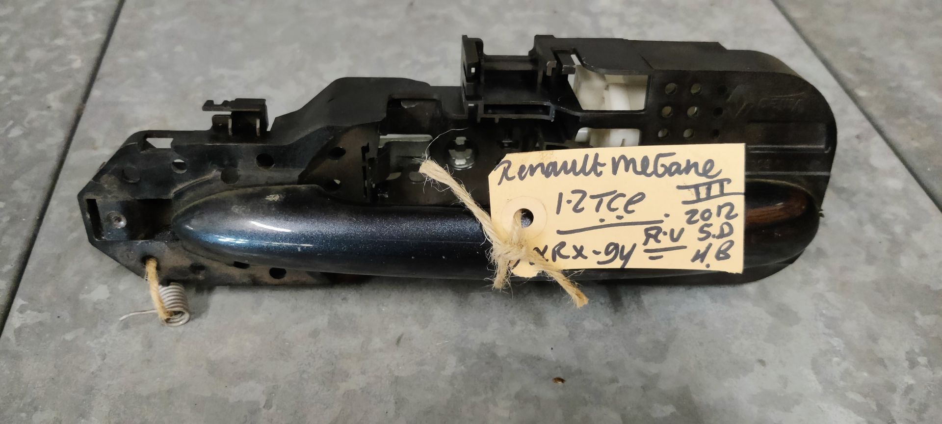 Een zwarte deurgreep voor een Renault Megane III met een handgeschreven identificatielabel, bevestigd met een touwtje.
