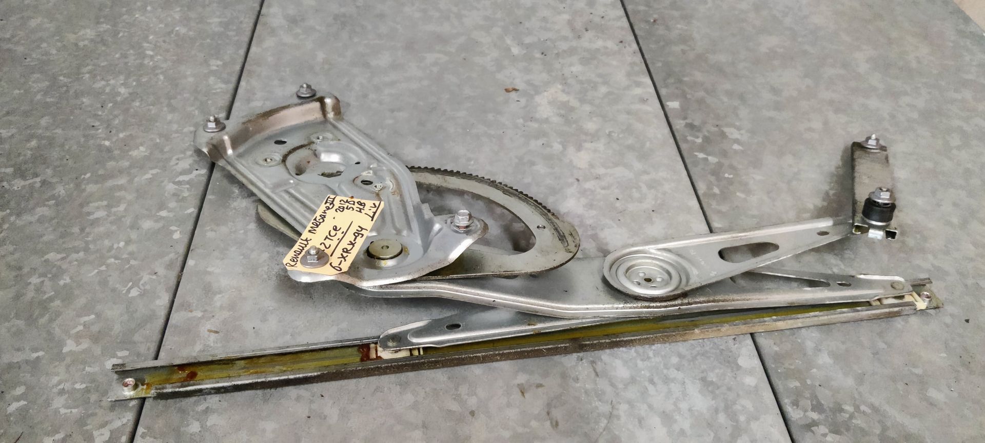 Een metalen raammechanisme voor een elektrische autoruit, met een tandwielmechanisme en een montagerail, op een betonnen vloer.