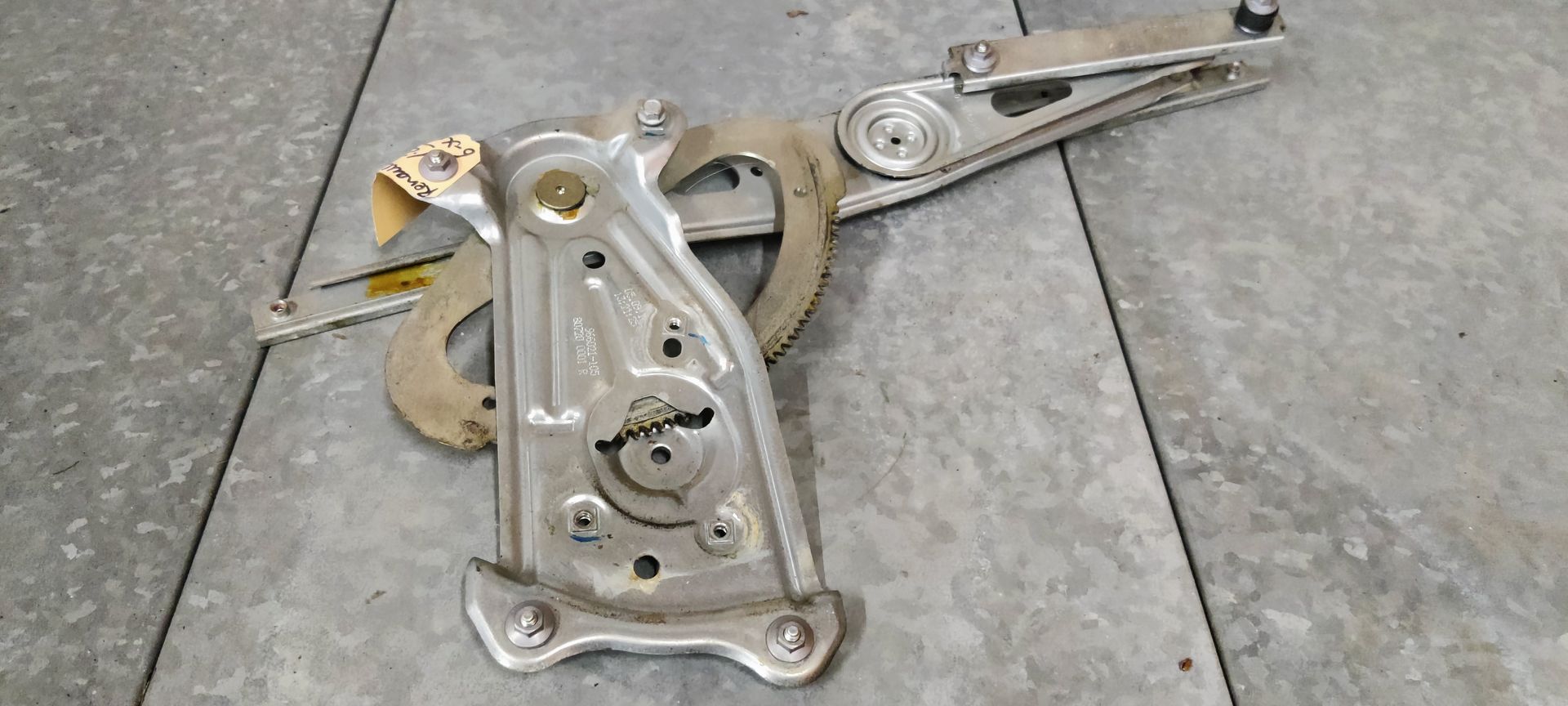 Een metalen raammechanisme van een auto ligt op een grijze tegelvloer.