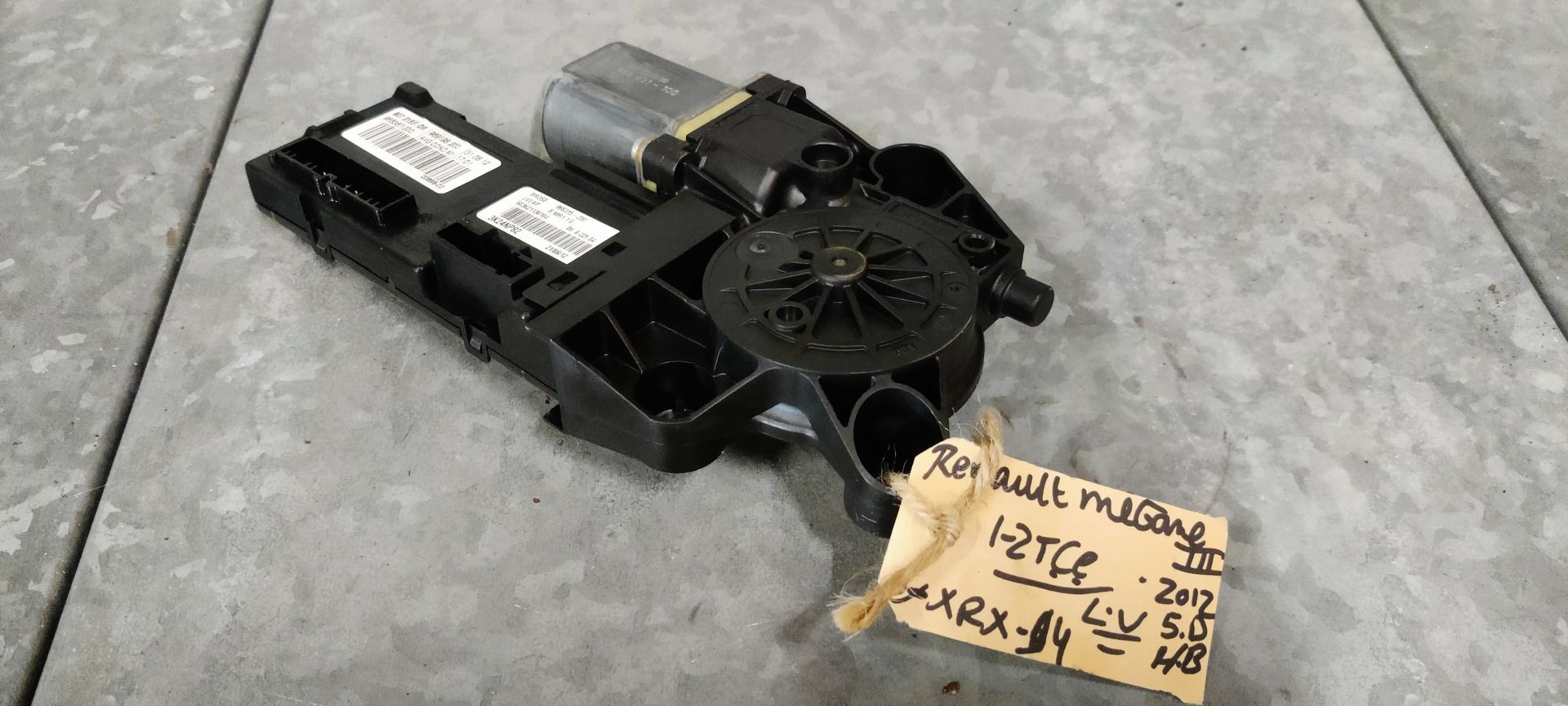 Een raammechanisme van een auto met een aangehechte zwarte plastic behuizing en een handgeschreven identificatielabel op een grijze vloer.