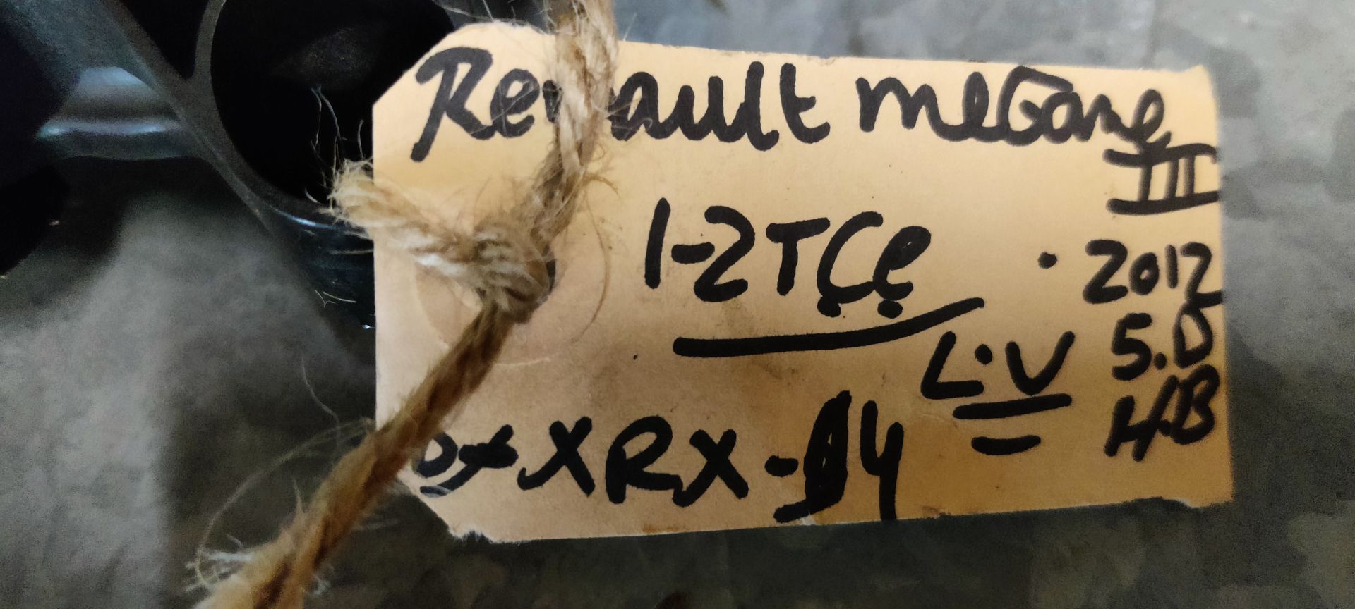 Een raammechanisme van een auto met een aangehechte zwarte plastic behuizing en een handgeschreven identificatielabel op een grijze vloer.