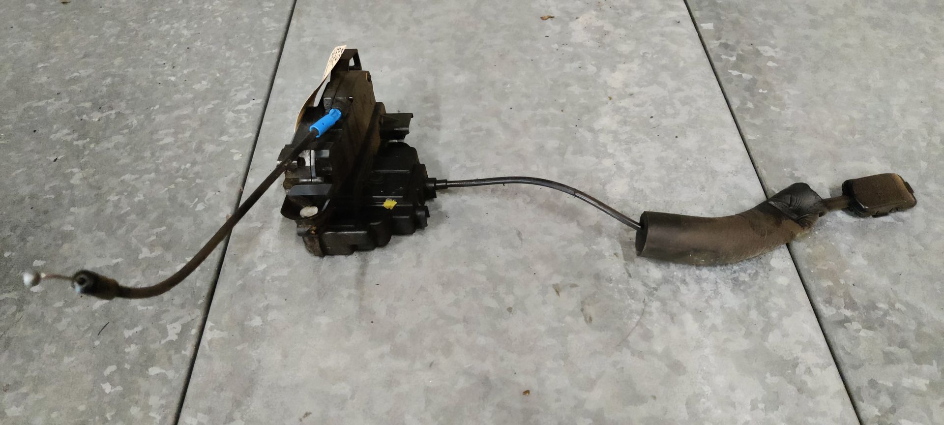 Een zwart autodeurslot met een aangehechte ontgrendelingskabel en beschermhuls, rustend op een grijze tegelondergrond.